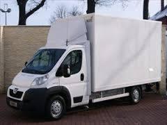 Biały bus dostawczy Peugeot z zabudową kontenerową, zaparkowany na brukowanym podjeździe, widok z boku, drzewo bez liści w tle.