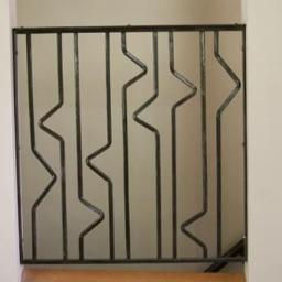 PW-POLMAR Sp. z o.o. - Metalowa balustrada schodowa z geometrycznym wzorem, zamontowana między białymi filarami. Ciemna konstrukcja na jasnym tle. Widok z bliska.