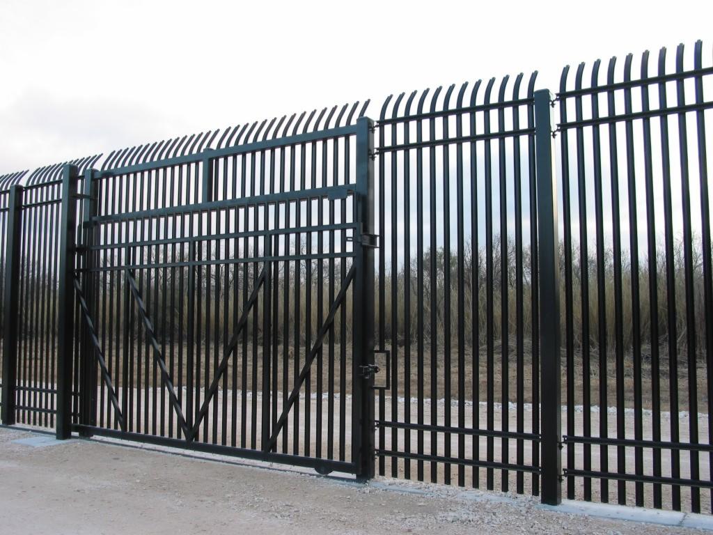 Czarne, metalowe ogrodzenie z bramą, pionowe pręty zakończone łukami, szary grunt, w tle roślinność. Solidna konstrukcja, ochrona posesji, nowoczesny design.