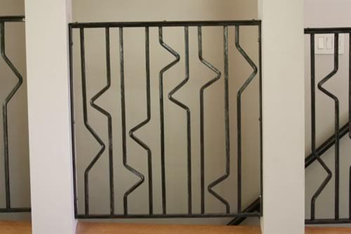 Metalowa balustrada schodowa z geometrycznym wzorem, zamontowana między białymi filarami. Ciemna konstrukcja na jasnym tle. Widok z bliska.