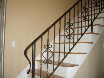 Kuta balustrada schodowa z dekoracyjnymi elementami w kształcie rombów i łuków, czarna, z delikatnymi złotymi akcentami, na tle beżowej ściany i białych stopni.