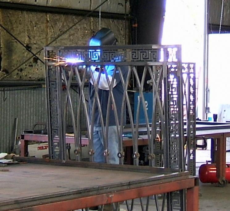 Spawanie metalowej konstrukcji z motywem greckim przez spawacza w masce. Iskry, warsztat, metalowy stół, częściowo widoczny zbiornik.