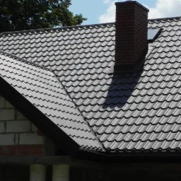 Nowy, brązowy dach z blachy falistej i komin z cegły na tle błękitnego nieba w Lublinie. Widoczne fragmenty murów budynku w stanie surowym.