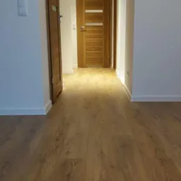 Korytarz z nową, jasną podłogą drewnianą ułożoną wzdłuż, widoczne dwie drewniane drzwi z przeszkleniami i białe listwy przypodłogowe.