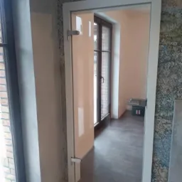Szklane drzwi wewnętrzne z białym obramowaniem i metalowymi okuciami, widoczne z boku, z oknem w tle i fragmentem ściany wykończonej płytkami imitującymi kamień.