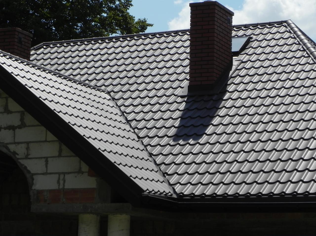 Nowy, brązowy dach z blachy falistej i komin z cegły na tle błękitnego nieba w Lublinie. Widoczne fragmenty murów budynku w stanie surowym.