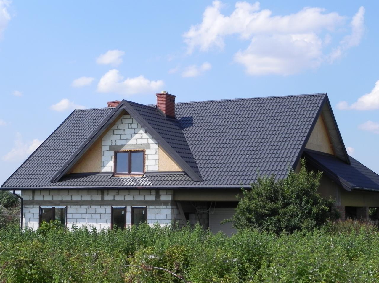 Dom w budowie z ciemną dachówką na dachu, widoczne okna i komin z cegły. Budynek otoczony zielenią, niebo błękitne z chmurami w tle.