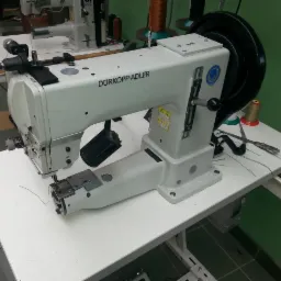 Profesjonalna maszyna szwalnicza Dürkopp Adler na białym stole, z widocznymi narzędziami i akcesoriami, w warsztacie z zielonymi ścianami.