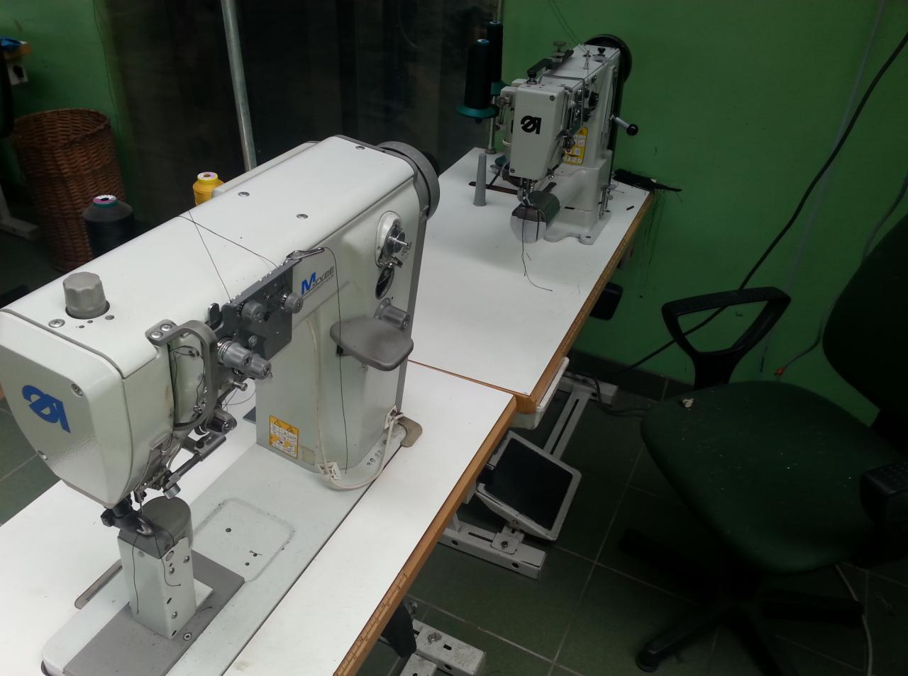 Dwie białe maszyny do szycia na białym stole, w tle zielona ściana i krzesło. Jedna maszyna marki M-Type. Widoczne nici i elementy mechaniczne.
