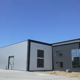 Nowoczesny budynek przemysłowy z szarej blachy trapezowej i ciemnoszarego tynku, duże okna, błękitne niebo, kostka brukowa, widok z zewnątrz.