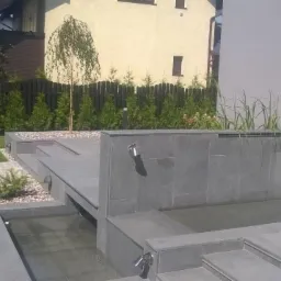 Nowoczesny ogród z kaskadą wodną wyłożoną szarą płytką, obok zadbany trawnik i tuje przy drewnianym płocie.