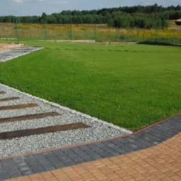 Ogród z równo skoszoną trawą, ścieżką z drewnianych elementów i kamienia, oraz altaną z trzciny w tle. Widok na zadbany teren zielony.