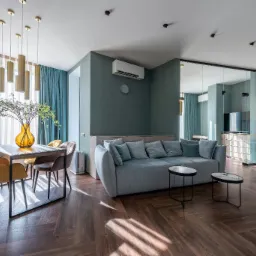 Nowoczesne wnętrze salonu z klimatyzacją ścienną, drewnianą podłogą w jodełkę, turkusowymi zasłonami i szklaną ścianką działową.