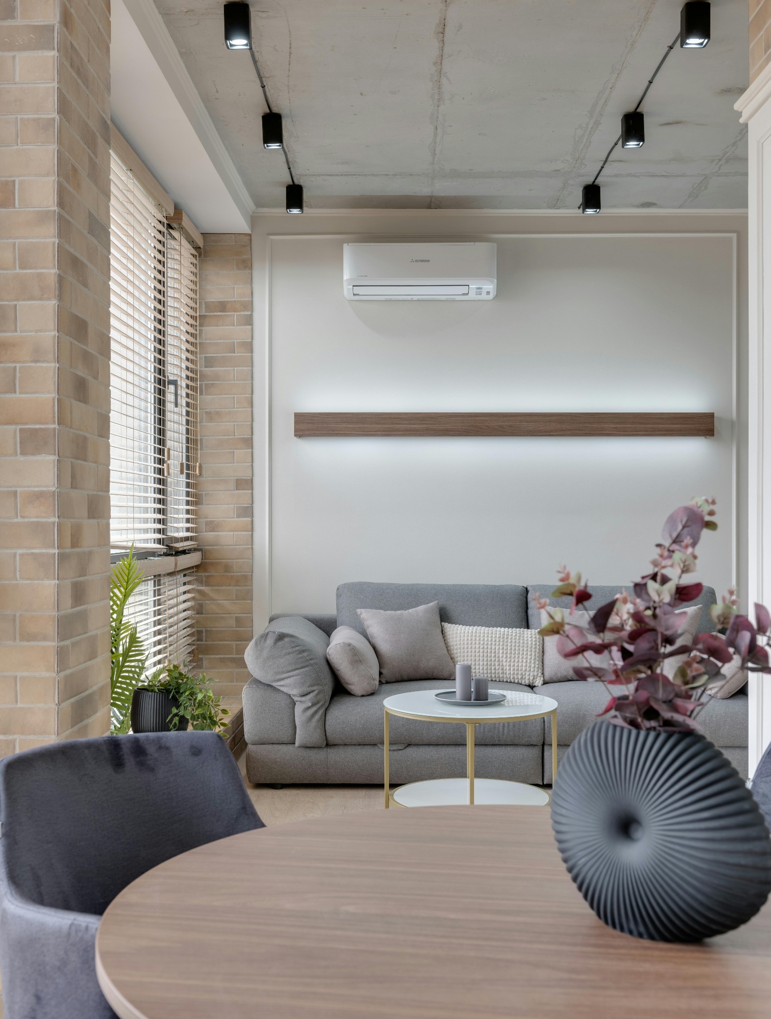 Wnętrze salonu z zamontowaną klimatyzacją Mitsubishi, widoczną na ścianie nad szarą sofą, ceglana ściana z oknem w tle, nowoczesne oświetlenie punktowe na betonowym suficie.