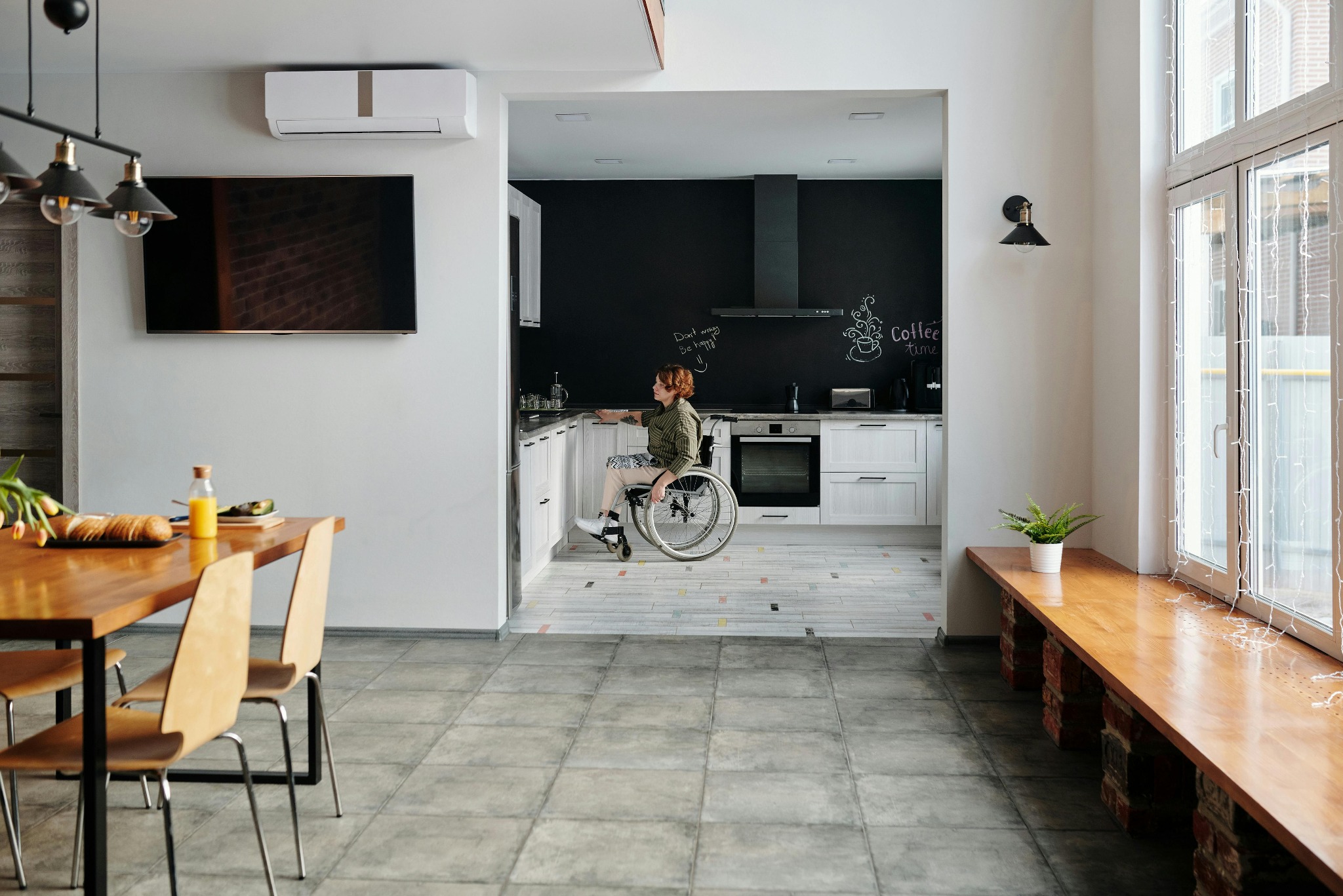 Wnętrze jasnego pomieszczenia z klimatyzatorem ściennym, widok na kuchnię z osobą na wózku inwalidzkim przy blacie.