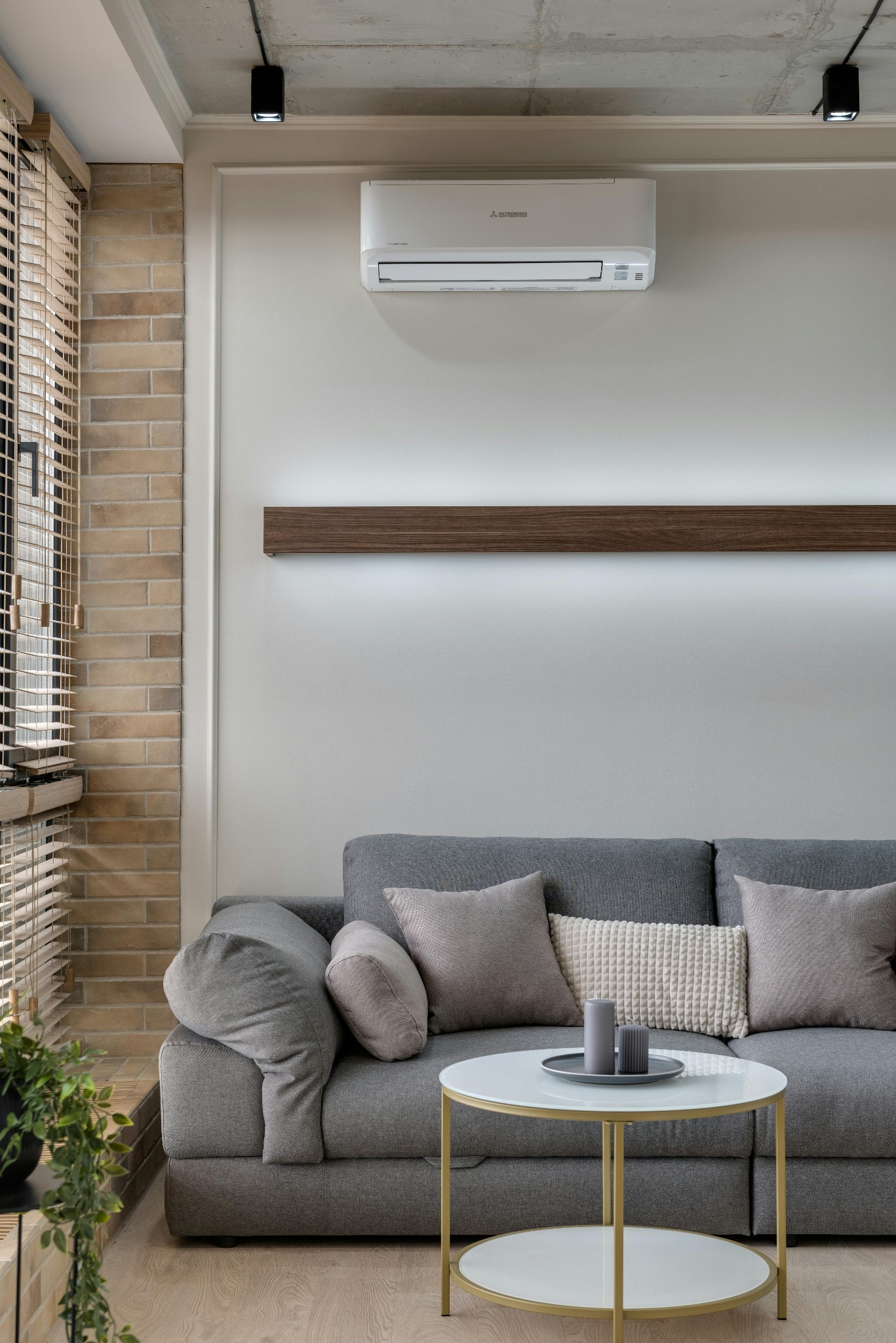 Biały klimatyzator Mitsubishi zamontowany na ścianie nad szarą sofą w nowoczesnym salonie z ceglaną ścianą i drewnianymi żaluzjami.