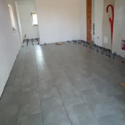 Szare płytki podłogowe ułożone w dużym pokoju, z systemem poziomowania płytek na krawędziach, widoczna instalacja grzewcza.