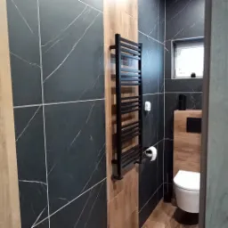 Nowoczesna łazienka z czarnymi płytkami imitującymi marmur, drewnopodobnymi akcentami i czarnym grzejnikiem drabinkowym; widoczny fragment toalety i okno z małą rośliną.