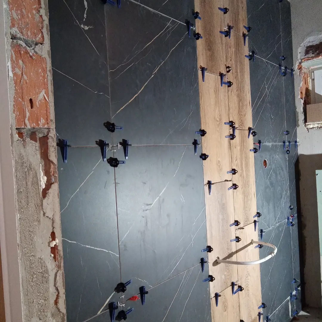 Ściana w trakcie układania płytek imitujących marmur i drewno, z użyciem systemu poziomowania płytek, widoczne surowe wykończenie ściany obok.