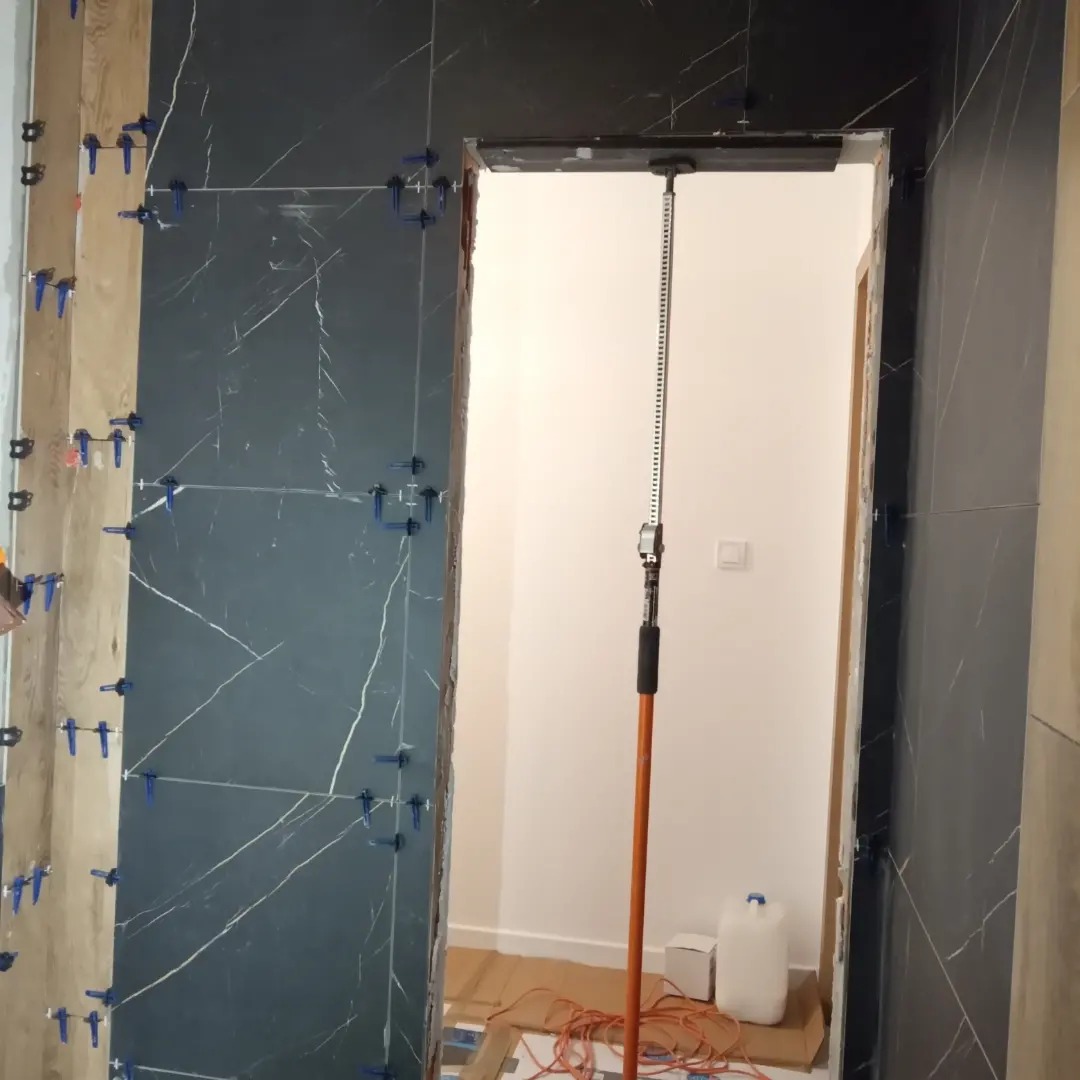 Montaż płytek imitujących marmur w ościeżnicy, z użyciem poziomicy laserowej na wysięgniku i klipsów poziomujących, widoczne przewody elektryczne na podłodze.