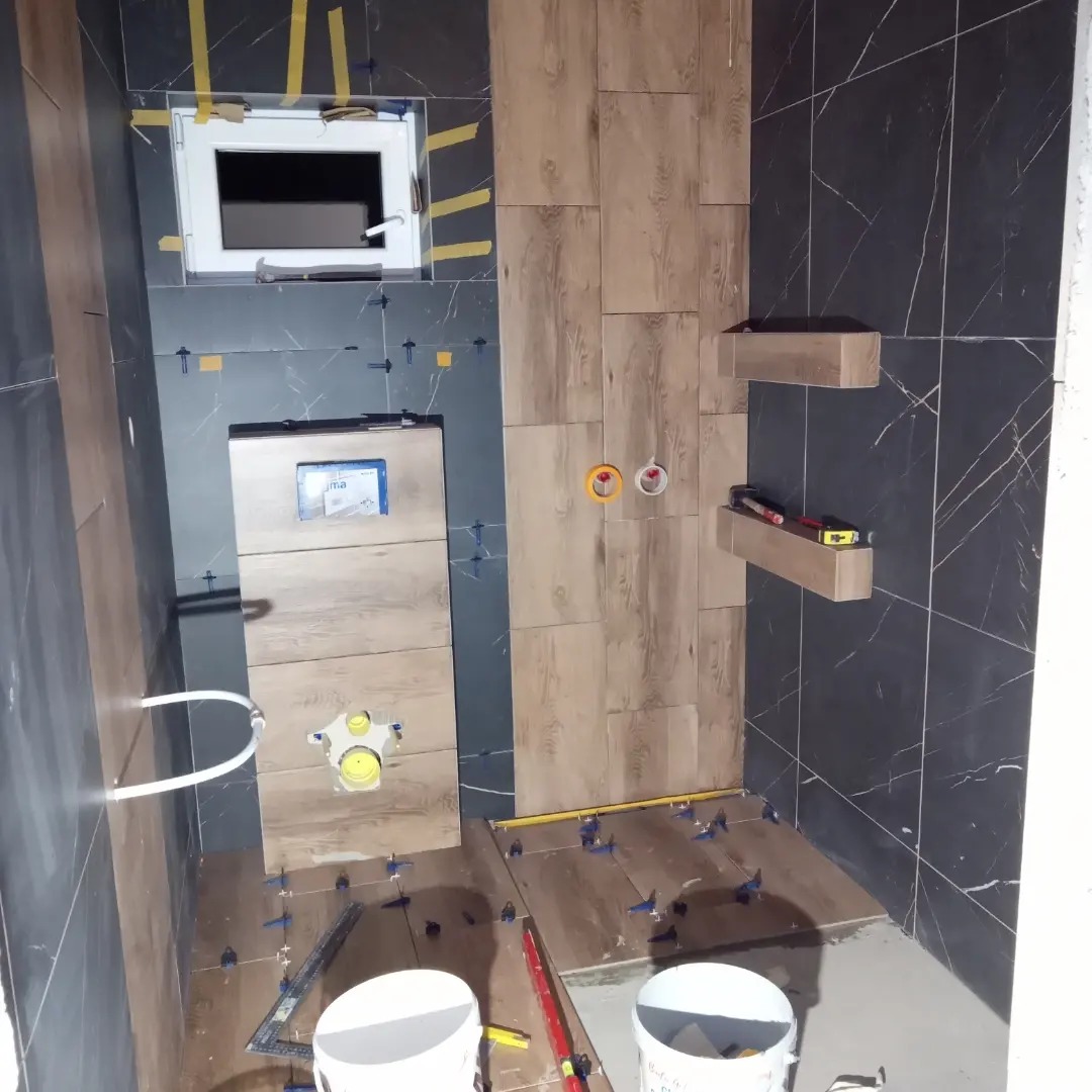 Łazienka w trakcie układania płytek imitujących drewno na podłodze i ścianie, widoczne krzyżyki dystansowe, dwa wiadra i poziomica, instalacja podtynkowa WC w trakcie montażu.