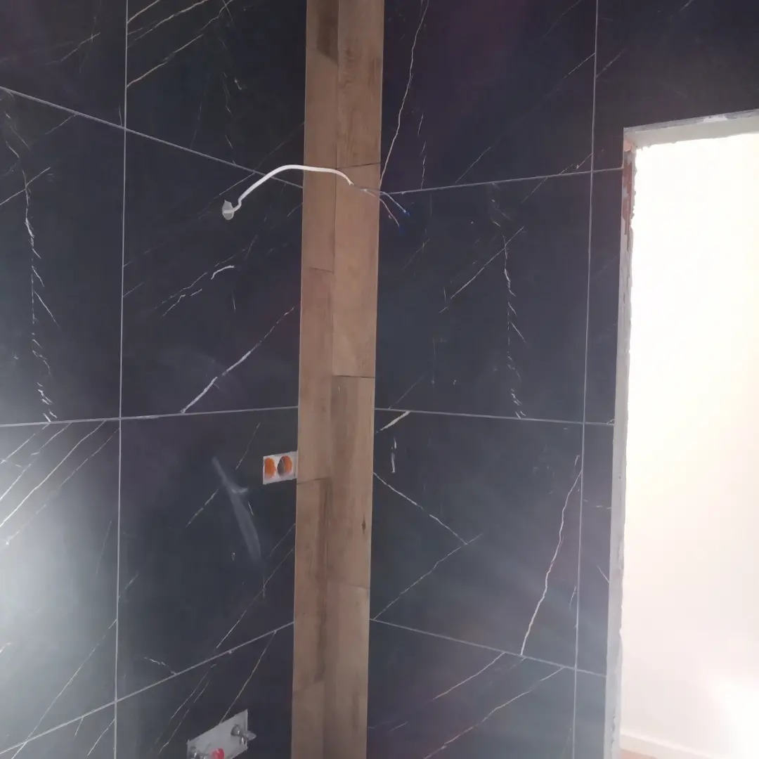 Częściowo wykończona łazienka z czarnymi płytkami marmurkowymi, drewnianą kolumną maskującą instalacje, wystającymi przewodami i gniazdkami elektrycznymi.