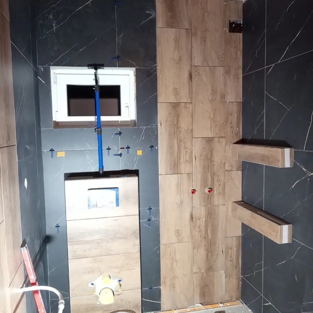 W trakcie montażu płytek imitujących drewno w łazience z ciemnymi płytkami marmurkowymi, widoczne poziomice i krzyżyki dystansowe.