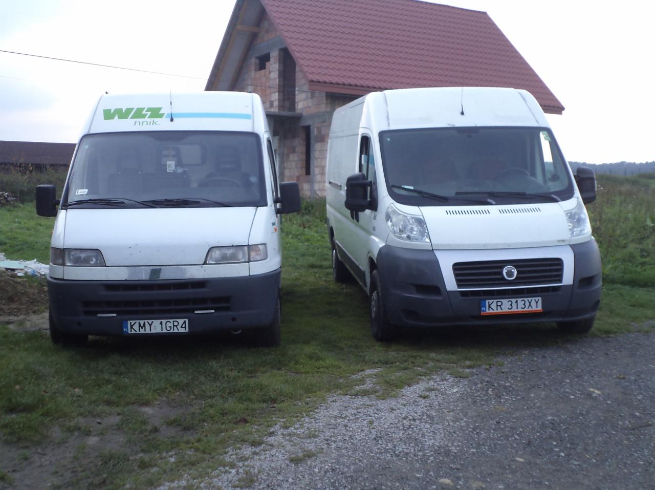 Dwa białe busy zaparkowane przed budynkiem w budowie; jeden z logo 'WIZZ nnik.', drugi z rejestracją 'KR 313XY'.