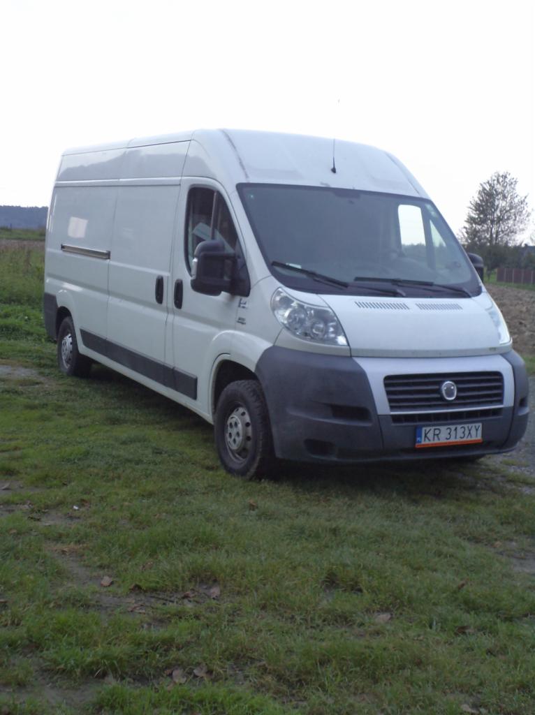 Biały bus dostawczy na trawie, z widoczną tablicą rejestracyjną KR 313XY i otoczeniem polnym w tle. Pojazd zaparkowany bokiem, z widoczną przednią częścią.