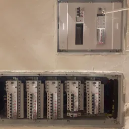 Dwa rozdzielnice elektryczne zamontowane w ścianie, widoczne kable i bezpieczniki, ściana w trakcie wykańczania.