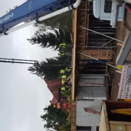 Wylewanie betonu za pomocą pompy na budowie domu jednorodzinnego; widoczne szalunki, pracownicy w kamizelkach odblaskowych oraz reklama budowy domów pod klucz.