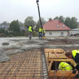 Wylewanie betonu na zbrojenie stropu budynku jednorodzinnego, widoczni pracownicy w żółtych kamizelkach, pompa do betonu, szalunki, w tle zabudowa jednorodzinna w pochmurny dzień.