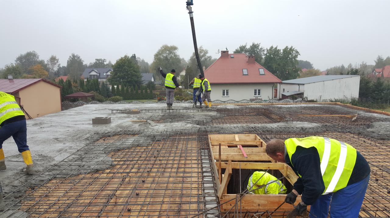 Wylewanie betonu na zbrojenie stropu budynku jednorodzinnego, widoczni pracownicy w żółtych kamizelkach, pompa do betonu, szalunki, w tle zabudowa jednorodzinna w pochmurny dzień.