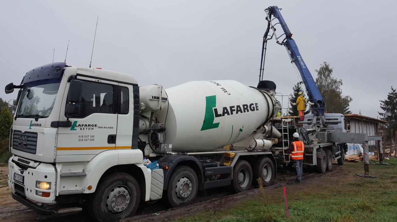 Betonomieszarka Lafarge z wysięgnikiem pompuje beton na budowę domu jednorodzinnego; widoczni pracownicy w kaskach i kamizelkach odblaskowych, szare niebo w tle.