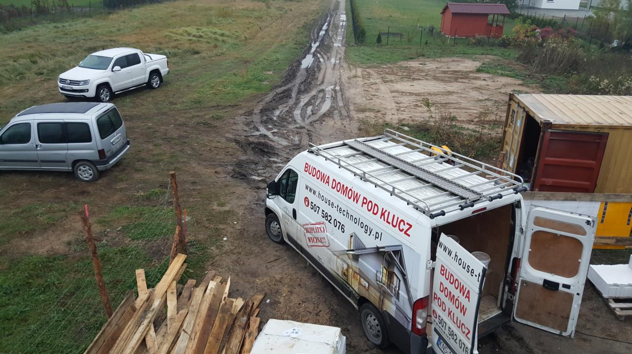 Plac budowy z błotnistą drogą, dwoma samochodami osobowymi, pickupem, kontenerami, stosem drewna i busem firmy budowlanej z otwartymi drzwiami, widok z góry.