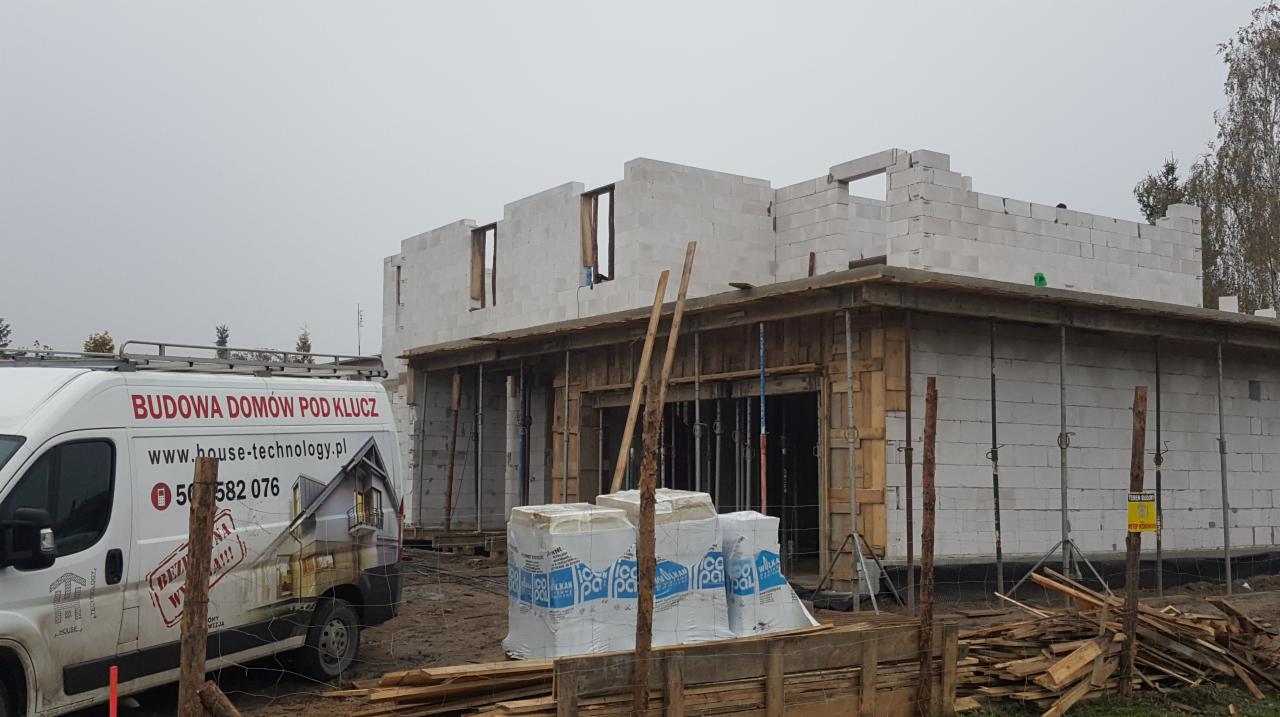 Budowa domu w trakcie wznoszenia ścian z betonu komórkowego, widoczny częściowo samochód dostawczy z reklamą firmy budowlanej na boku, oraz drewniane szalunki i podpory.