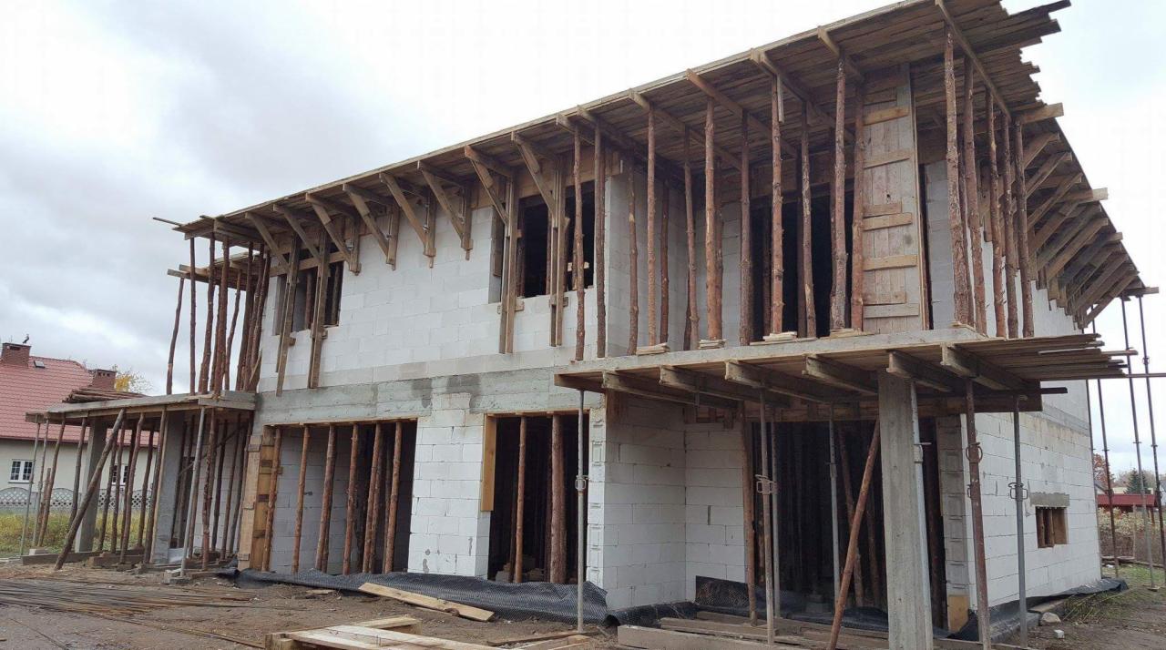 Dwukondygnacyjny dom w trakcie rozbudowy, ściany z białych bloczków, drewniane stemple podtrzymujące konstrukcję stropu i dachu, widoczne zbrojenia i szalunki.