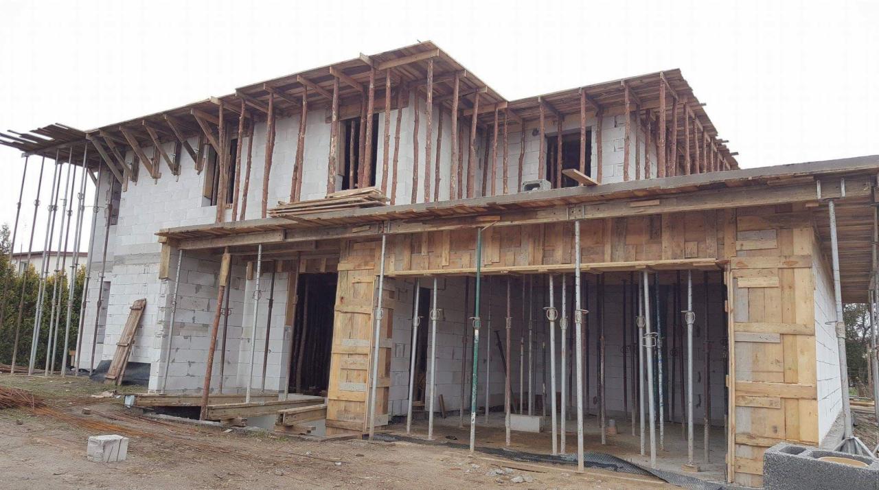 Surowy stan rozbudowy domu z białych bloczków, z widocznym drewnianym szalunkiem i metalowymi podporami, teren budowy z resztkami materiałów.