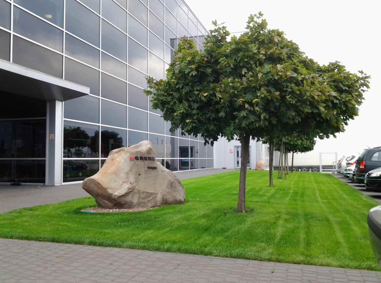 Zadbany trawnik z równo posadzonymi drzewami kulistymi przed nowoczesnym budynkiem z przeszkloną fasadą. Na trawniku duży kamień z napisem.