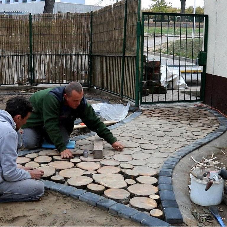 Dwóch mężczyzn układa ścieżkę ogrodową z drewnianych krążków i kostki brukowej, widoczny bambusowy płot i metalowa furtka.