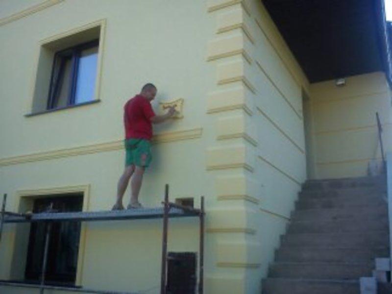 Pracownik na rusztowaniu maluje dekoracyjny element elewacji budynku w Łodzi. Jasna elewacja z żółtymi akcentami wokół okien i na narożnikach.