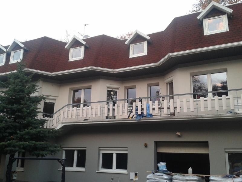 Dwukondygnacyjny dom z balkonem po odświeżeniu elewacji, widoczne worki z materiałem budowlanym i wiadro farby u dołu zdjęcia. Czerwony dach, jasne ściany.