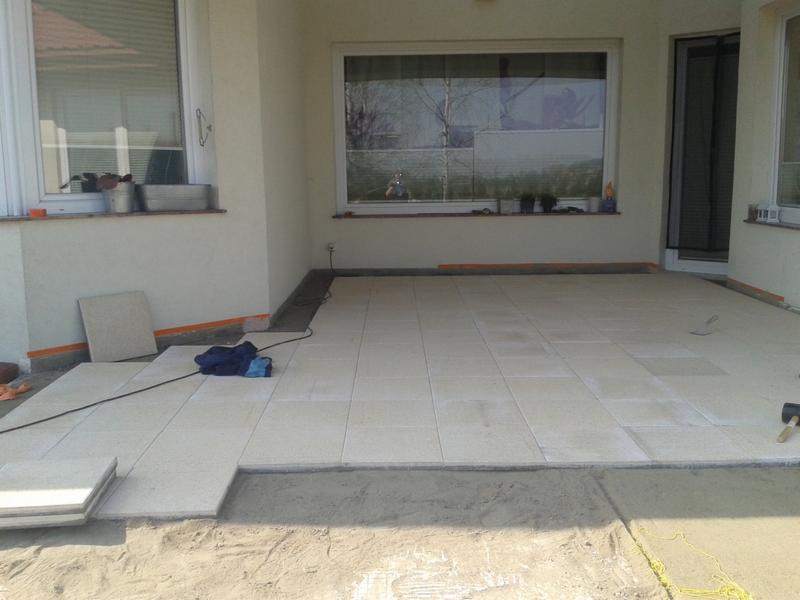 Układanie jasnych płytek na tarasie domu jednorodzinnego. Widoczne okna i drzwi balkonowe. Etap prac: przygotowanie podłoża i częściowe ułożenie płytek.