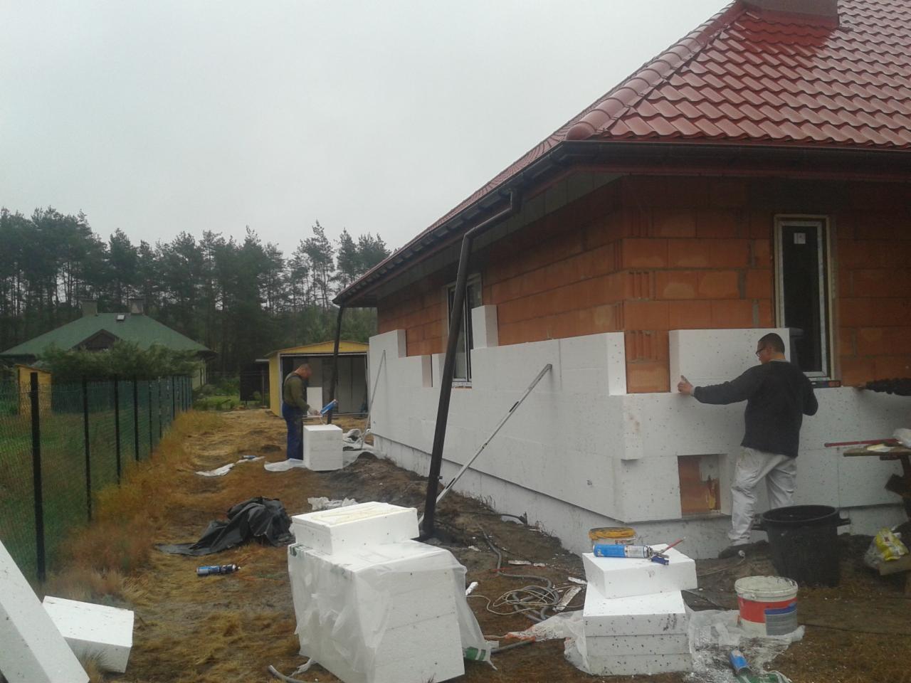 Ocieplanie domu styropianem: pracownicy montują białe płyty izolacyjne na elewacji z cegły. Widoczne narzędzia i materiały budowlane. Tło: las i pochmurne niebo.