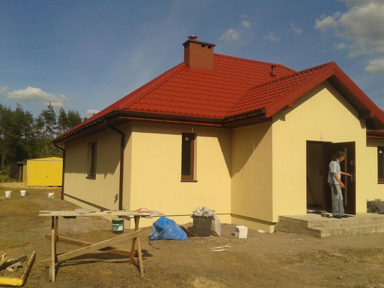 Żółty dom jednorodzinny z czerwoną dachówką po tynkowaniu, widoczny fragment rusztowania i osoba wchodząca do budynku. Słoneczny dzień.