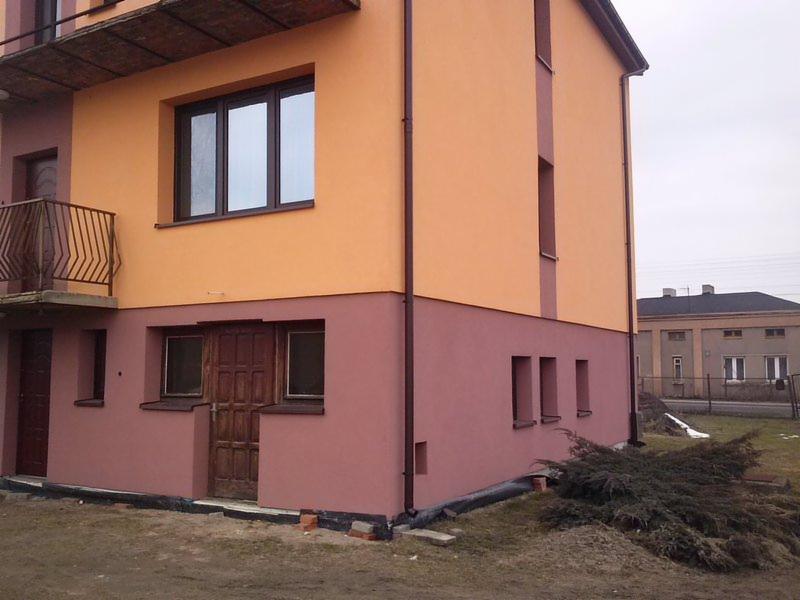 Dwukondygnacyjny budynek z elewacją w odcieniach pomarańczy i różu, z brązowymi oknami i drzwiami. Widoczne rury spustowe i fragment otoczenia z roślinnością.