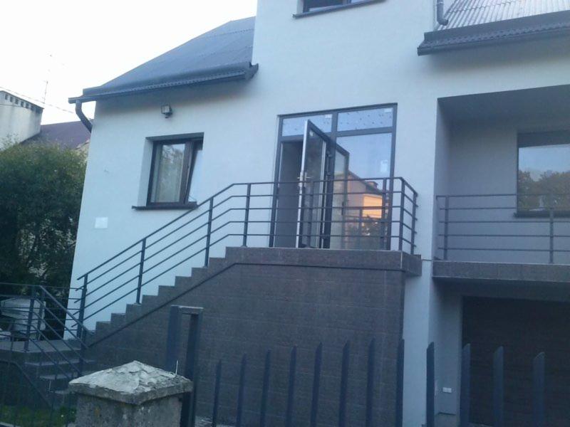 Elewacja domu po renowacji z grafitowymi detalami: schody z balustradą, okna i drzwi balkonowe. Jasna kolorystyka ścian i ciemny dach tworzą kontrast.