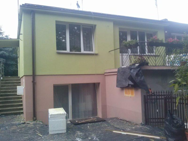 Dwukondygnacyjny dom z zieloną i różową elewacją. Część elewacji zakryta czarną folią. Widoczne okna, balkon z kwiatami i schody zewnętrzne.