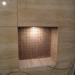 Wnęka w ścianie wyłożona beżowymi płytkami z dekoracyjnym oświetleniem punktowym i mozaiką z drobnych, brązowych płytek, wykończona listwą imitującą drewno.