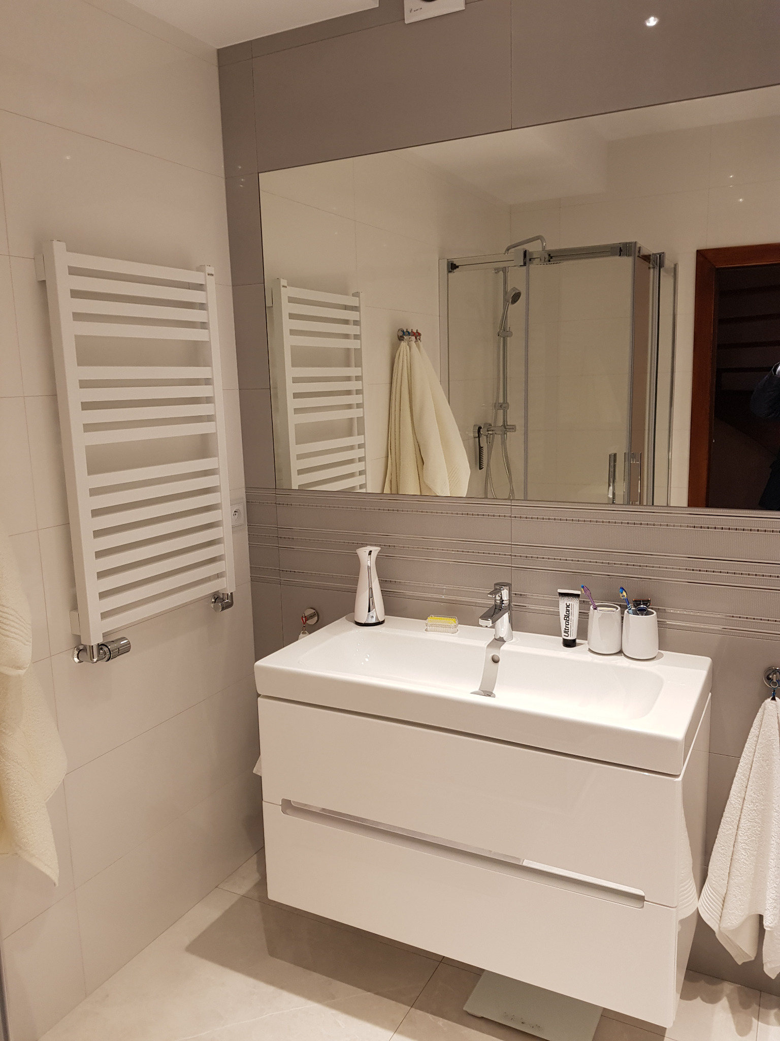 Nowoczesna łazienka z białą szafką umywalkową, grzejnikiem drabinkowym i dużym lustrem, w którym odbija się kabina prysznicowa i wiszący ręcznik.